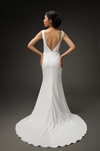 A young woman wearing a sleek, white wedding dress with a deep V-back and a fitted silhouette that flows into a small train. She stands with her hands on her hips, showcasing the elegant lines of the dress from behind. Her hair is styled in an updo, and she wears delicate earrings. The dark background accentuates the dress's sophisticated design and her graceful posture.