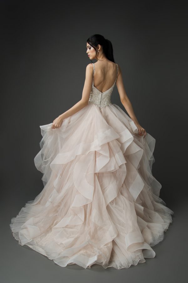 A woman wearing a blush organza wedding dress, viewed from the back. The dress features a fitted beaded bodice with thin straps and an open back, leading to a voluminous layered organza skirt in a soft blush color. The cascading ruffles of the skirt create a dramatic and flowing effect, enhancing the romantic and elegant look of the gown.