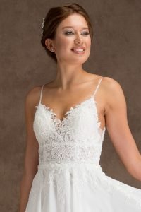 A close-up of a young woman wearing the "Lacy" beaded chiffon wedding dress, showcasing the intricate lace bodice and thin straps. She smiles softly, with her hair styled up and a decorative hairpiece. The plain brown background highlights her elegant appearance.