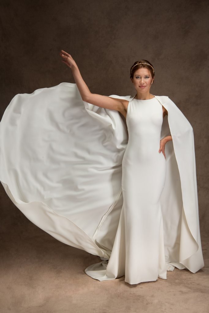 A woman poses in a sleek white bridal gown with a high neckline and flowing train. She wears a dramatic bridal cape billowing behind her and a pearl-accented headband, adding a regal touch to her elegant look.