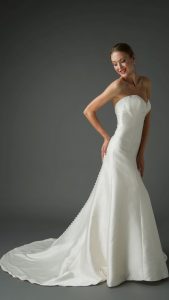 A young woman wearing a strapless trumpet wedding dress that elegantly hugs her figure and flares out at the bottom. The dress features a smooth, satin finish with a row of buttons running down the back. She poses with a smile, hands on her hips, and her hair styled in an elegant updo.