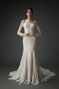 Bride in an art deco beaded wedding dress with intricate detailing, long sheer sleeves, and a dramatic mermaid silhouette, standing against a dark background.