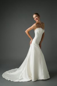 A young woman wearing a strapless trumpet wedding dress that elegantly hugs her figure and flares out at the bottom. The dress features a smooth, satin finish with a row of buttons running down the back. She poses with a smile, hands on her hips, and her hair styled in an elegant updo. The dark background highlights the sleek lines and sophisticated design of the dress.