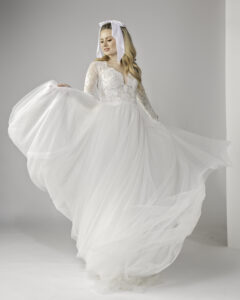 Flowing long sleeve lace and tulle wedding dress with a lace bodice and soft, airy skirt. The bride gracefully lifts the skirt, showing off the dress's movement, while a large white bow in her hair adds a charming, vintage-inspired accent to her look.