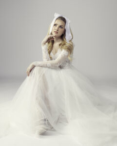 Whimsical long sleeve lace and tulle wedding dress, featuring a delicate lace bodice and flowing tulle skirt. The bride sits thoughtfully, wearing a large white bow in her hair, adding a playful and vintage-inspired touch to her bridal look.