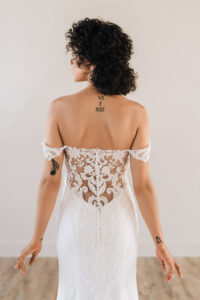 A woman wearing a lace off-the-shoulder wedding dress. The dress features intricate lace detailing along the back, with delicate buttons running down the center. The off-the-shoulder sleeves add a romantic and elegant touch, complementing the fitted silhouette of the gown. The lace pattern is beautifully designed to highlight the curves, and the sheer elements add sophistication to the overall look.