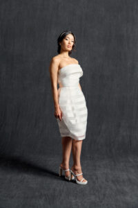 Asian woman in a strapless short satin wedding dress with tiered layers, standing in a poised pose with white high-heeled sandals, against a dark, textured backdrop.