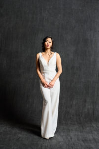 A bride poses confidently in a modern bridal jumpsuit featuring a deep V-neckline and a fitted waist adorned with delicate beadwork. The wide-leg pants create a sleek and elegant silhouette, perfectly complementing the minimalist design. Her dark, wavy hair is styled in loose curls, adding a touch of softness to her sophisticated look.