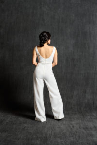 A back view of a modern bridal jumpsuit. The jumpsuit features a deep V-back, accentuated with delicate beadwork along the neckline and waistline. The fabric drapes elegantly, highlighting the bride's silhouette with its tailored fit and flared legs. The bride's hair is styled in a chic updo, completing the contemporary bridal look.