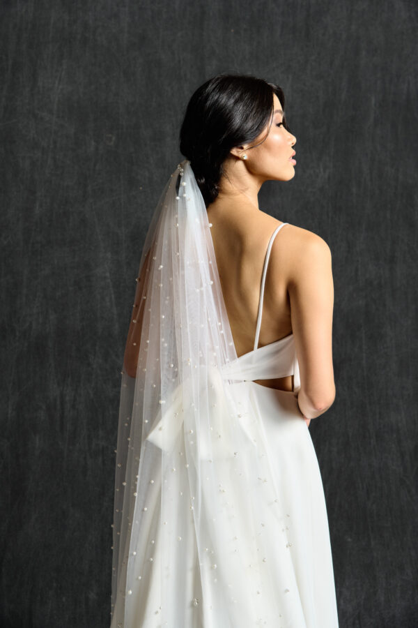A bride stands gracefully in profile, showcasing a delicate, pearl-adorned veil that cascades down her back. The veil is attached to her dark, elegantly styled hair, which complements her simple yet sophisticated wedding dress with thin straps and a low back. The dark background contrasts with the soft white of her veil and dress, highlighting the intricate pearl details and the elegant simplicity of her bridal look.