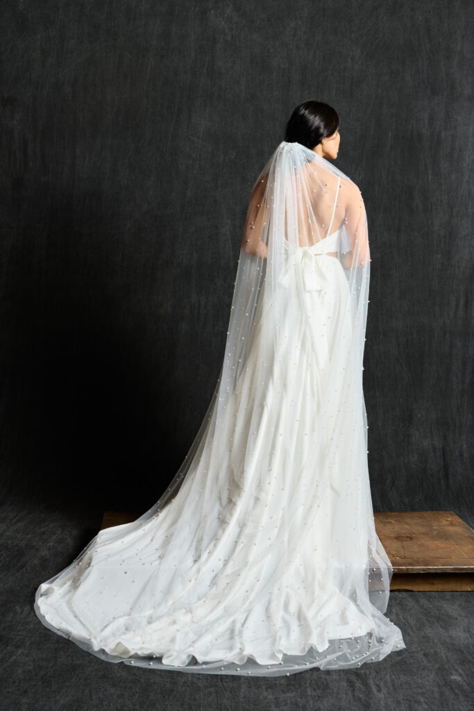 A bride stands with her back to the camera, showcasing a long, flowing wedding dress and a delicate veil. The dress features a soft, draping fabric that creates an elegant train on the floor. The veil is adorned with small, scattered pearls, adding a touch of sparkle and sophistication. The dark background contrasts with the white dress and veil, emphasizing their intricate details. The bride's hair is styled in a simple, elegant manner, completing the refined and graceful look.