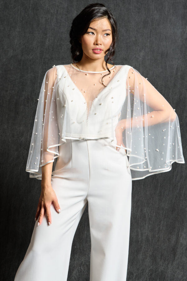 A bride is shown wearing a bridal jumpsuit paired with a delicate beaded capelet. The capelet, made from sheer fabric, is adorned with pearls, adding a soft, romantic touch to the modern, structured outfit. The combination of the sleek jumpsuit and the detailed capelet creates a unique and contemporary bridal look.