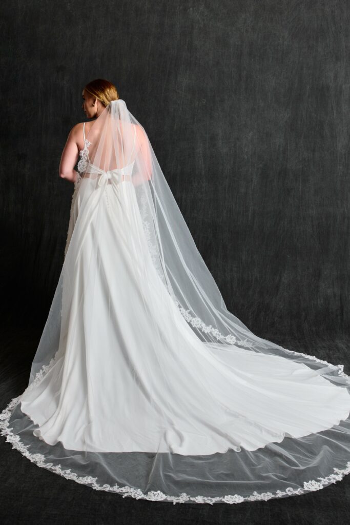 A young woman wearing a beaded chiffon wedding dress with a lace bodice and thin straps. She holds the sides of the flowing skirt, smiling brightly. Her hair is styled up with a decorative hairpiece, and the plain brown background accentuates the elegance of the dress and her joyful expression. She is also wearing a cathedral veil with a lace edge.