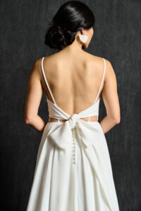 A close-up view of the back of a modern bridal gown featuring delicate spaghetti straps and an open back. The dress is adorned with a large, elegant bow at the waist, adding a touch of femininity and charm. Small buttons trail down from the bow, adding detail and sophistication. The bride's hair is styled in a low, elegant bun, and she wears statement earrings that complement the minimalist yet refined design of the gown.