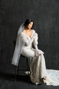 A bride sits gracefully on a stool, wearing an intricately designed lace wedding gown with long sleeves and a deep V-neckline. The dress features a vintage-inspired pattern, adding a touch of elegance and sophistication. Her sheer veil falls gently over her shoulders, complementing the dress's detailed lacework. The backdrop is a dark, textured surface that highlights the delicate lace and the flowing train of the gown. The bride's pose and serene expression enhance the timeless beauty of the ensemble.