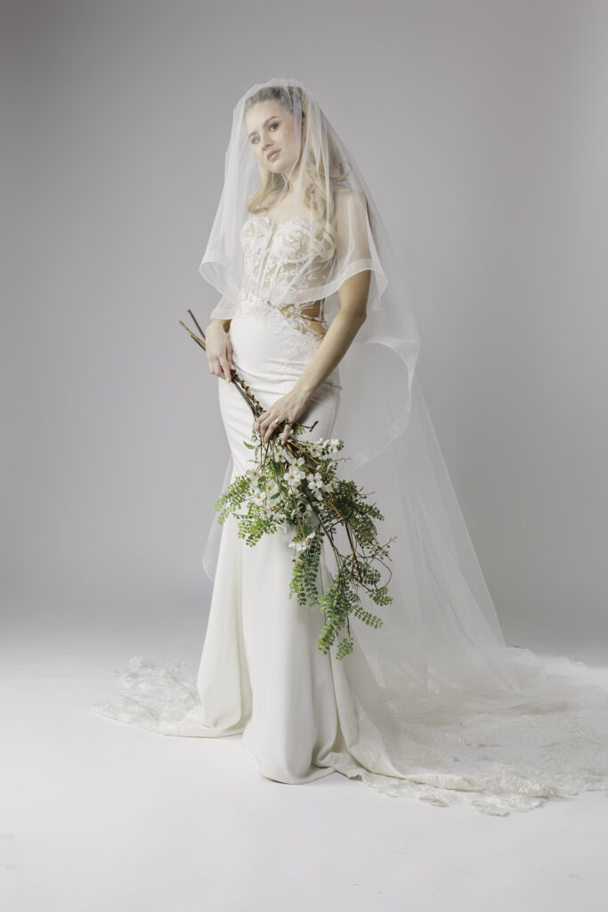 A lace corset wedding dress with a long veil. The dress features a structured lace bodice with visible corset boning and a fitted, smooth skirt that extends into a lace train. The woman holds a bouquet of wild branches and greenery, adding a natural, rustic touch to her elegant look. The soft, sheer veil frames her face, enhancing the romantic feel of the ensemble.