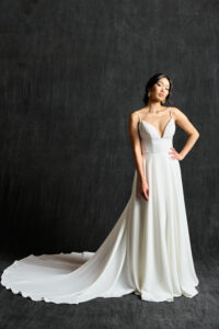 A modern bridal gown featuring delicate spaghetti straps and a plunging V-neckline. The dress has a fitted bodice that flows into a voluminous, elegant skirt with a long train. The clean, minimalist design highlights the bride's natural beauty and grace. The bride completes the look with simple, chic earrings, and her hair is styled in a sophisticated updo.
