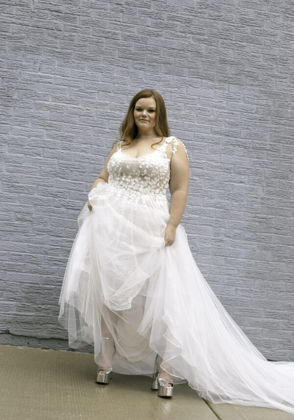 Woman in a 3D floral wedding dress with a bodice covered in floral appliqués and a layered tulle skirt, standing against a light gray brick wall. The dress's textured floral details and soft flow create an elegant and romantic bridal look.