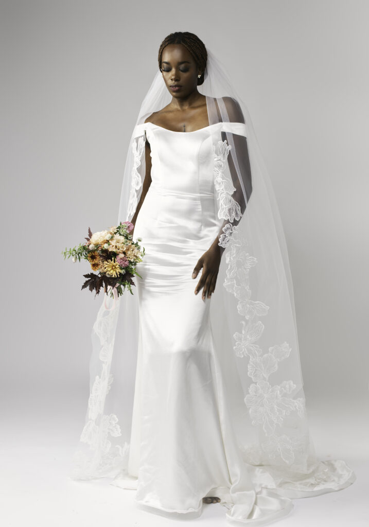Woman in an off-shoulder wedding dress holding a bouquet, wearing a chapel length lace cathedral veil.