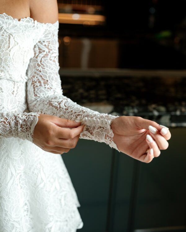Close-up of a bride's hand fastening the button on the lace sleeve of her off-the-shoulder wedding dress, showcasing the intricate lace detailing and her manicured nails.