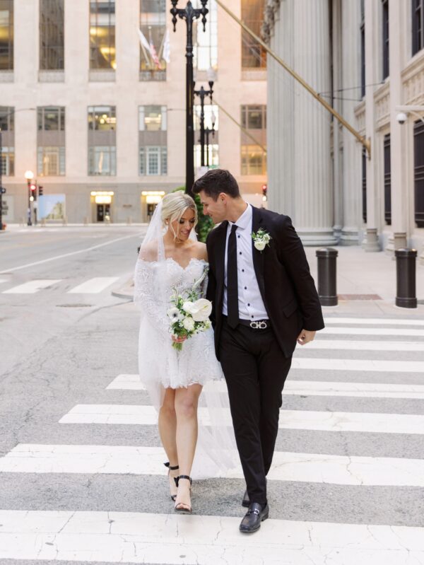 Bride and groom walking hand-in-hand across a city crosswalk, with the bride wearing a short lace wedding dress, a veil, and carrying a bouquet of white flowers. The groom is dressed in a black suit with a boutonnière.