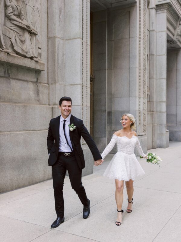 Bride and groom joyfully walking hand-in-hand outside, with the bride wearing a short lace wedding dress and a bouquet of white flowers. The groom is dressed in a black suit with a white boutonnière.