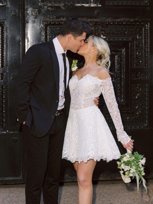 Bride and groom sharing a kiss in front of a dark door. The bride is wearing a short lace wedding dress with long sleeves and a bouquet of white flowers.
