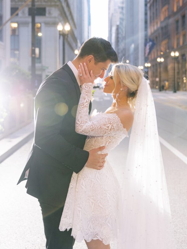 Bride and groom embracing in a city street, sharing a tender moment. The bride wears a short lace wedding dress with off-the-shoulder sleeves and a pearl-detailed veil.