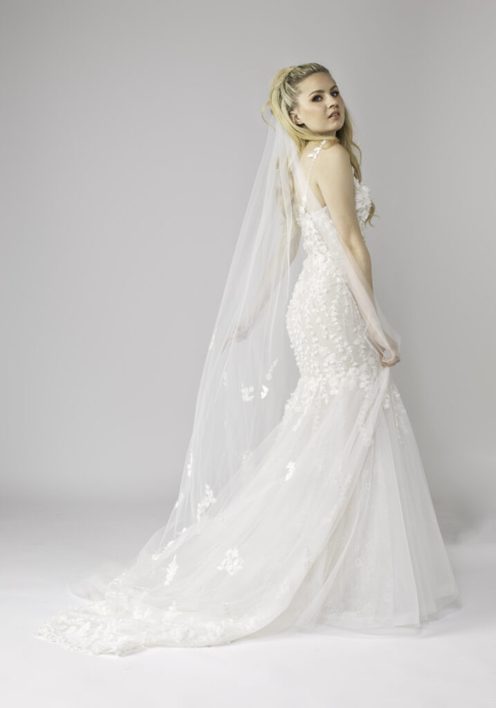 Woman in a lace wedding dress with a long 3D floral veil, looking over her shoulder.