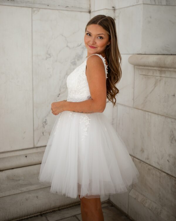 A smiling woman poses in a lace and tulle short wedding dress with a flared skirt and floral lace details, perfect for bridal celebrations.